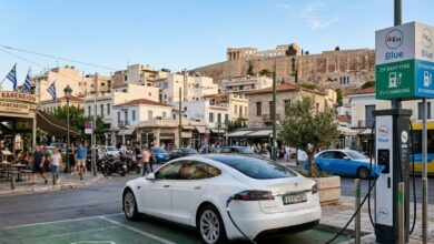 Σύγχρονο ηλεκτρικό αυτοκίνητο που φορτίζει σε δημόσιο σταθμό με φόντο ελληνικό αστικό τοπίο